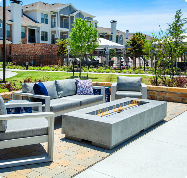 An outdoor lounge seating area with a modern rectangle-shaped glass fire pit surrounded by grassy lawn, trees and lush landscape at MAA Copper Ridge luxury apartment in Roanoke, TX