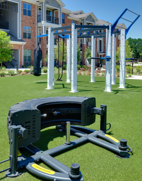 An outdoor workout station with a punching bag and climbing gym next to a tire flip training machine on turf grass surrounded by trees and plants at MAA Copper Ridge luxury apartments in Roanoke, TX