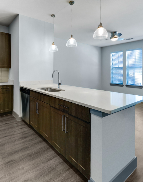 A contemporary kitchen with hanging light fixtures, dark wood cabinets with white granite countertops and wood flooring next to a living room space at MAA Copper Ridge luxury apartments in Roanoke, TX