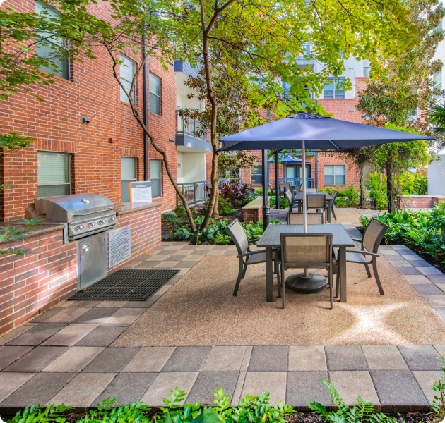 A modern outdoor patio area with an outdoor grill station, patio table with chairs and a large umbrella surrounded by greenery at MAA Eastside luxury apartments in Richardson, TX