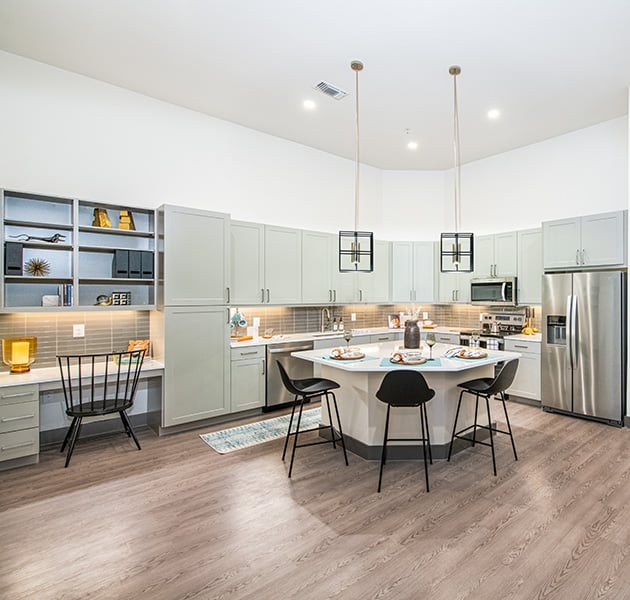 Open apartment kitchen with light grey cabinets, granite countertops, stainless steel appliances with tile backsplash, kitchen island countertop dining area with bar stools on wood floors at MAA Frisco Bridges luxury apartments in Frisco, TX