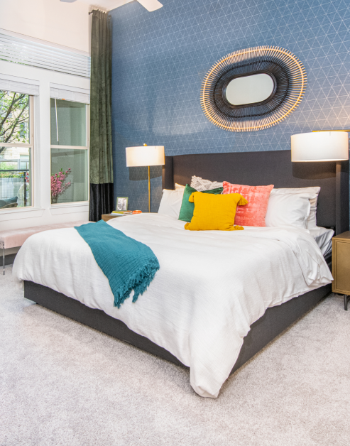 Model carpeted bedroom with accent blue wall with art and curtains and white bedding with accent pillows at MAA Frisco Bridges luxury apartments in Frisco, TX