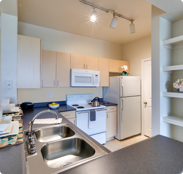 A light-colored kitchen with track ceiling lights and light wood cabinets with modern appliances and stainless steel double-sink next to a built-in shelf countertop at MAA Grand Courtyards luxury apartments in Grand Prairie, TX