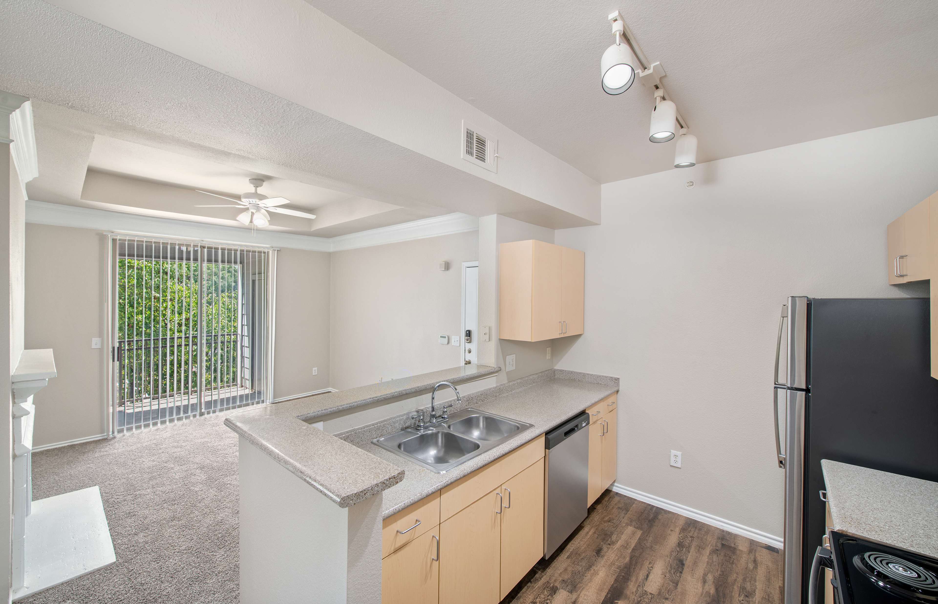 An outward view from kitchen features track ceiling lights over bar top counter and light tan wood cabinets positioned next to the living room with sliding glass doors at MAA Grand Courtyards luxury apartments in Grand Prairie, TX