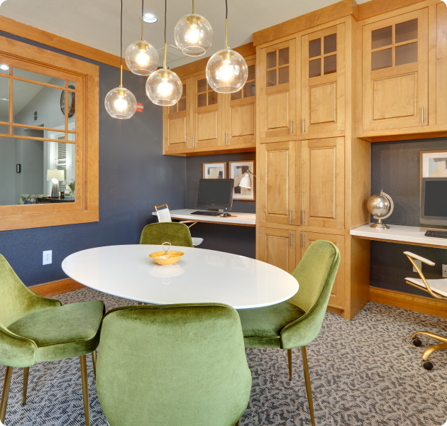 A upscale community work center with tan wall cabinetry, workstations and white oval table seating area with hanging globe light fixtures and green velvet chairs at MAA Grand Courtyards luxury apartments in Grand Prairie, TX
