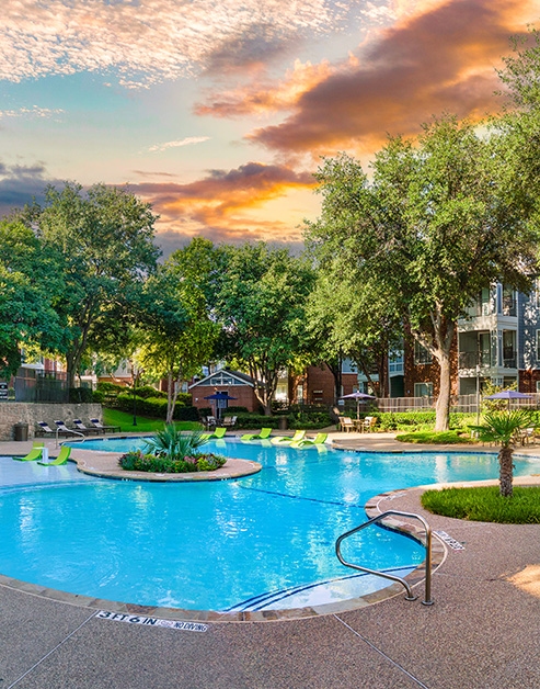 Resort-style swimming pool with a sundeck and island surrounded by trees at MAA Grand Courtyards luxury apartments in Grand Prairie, TX