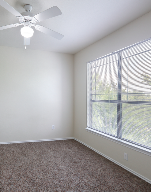 An open-space empty bedroom with a ceiling fan next to large window with blinds at MAA Grand Courtyards luxury apartments in Grand Prairie, TX