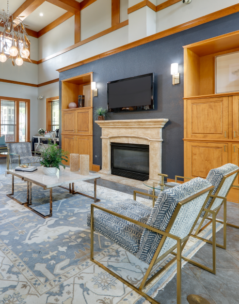 A luxurious community clubhouse features a wall-mounted TV above a stone fireplace with sleek wooden shelving next to sitting area with cushioned armchairs at MAA Grand Courtyards luxury apartments in Grand Prairie, TX