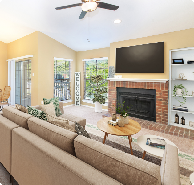 An apartment living room with vaulted ceilings and a ceiling fan, a wall-mounted TV above a fireplace next to sitting area with large windows with blinds and built-in shelves at MAA Grapevine luxury apartments in Grapevine, TX