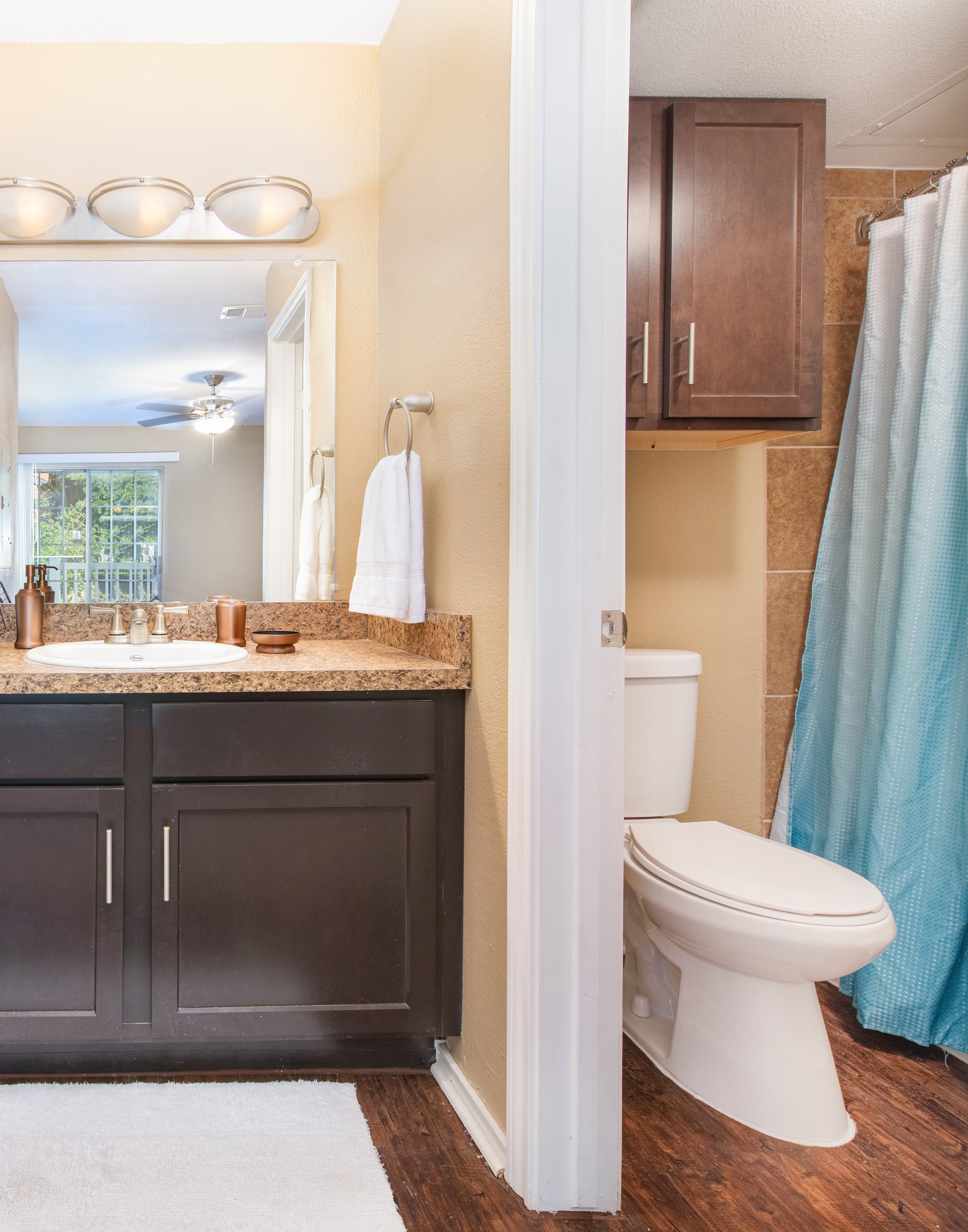 Bathroom with light fixtures above vanity mirror and granite countertop with single sink and dark brown cabinets, next to private toilet and shower area at MAA Grapevine luxury apartments in Grapevine, TX