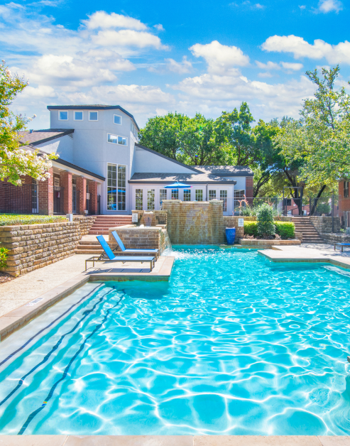 Side view of swimming pool with cascading waterfalls next to lounge chairs and steps to clubhouse, surrounded by trees at MAA Grapevine luxury apartments in Grapevine, TX