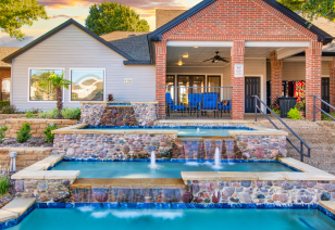 A poolside three-level waterfall fountain with small fountains sits next to a brick clubhouse with seating area at MAA Grapevine luxury apartments in Grapevine, TX