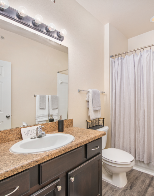 A modern bathroom featuring a large vanity mirror with lights, granite countertop and cabinets under the sink with hard flooring at MAA Hebron luxury apartments in Carrollton, TX