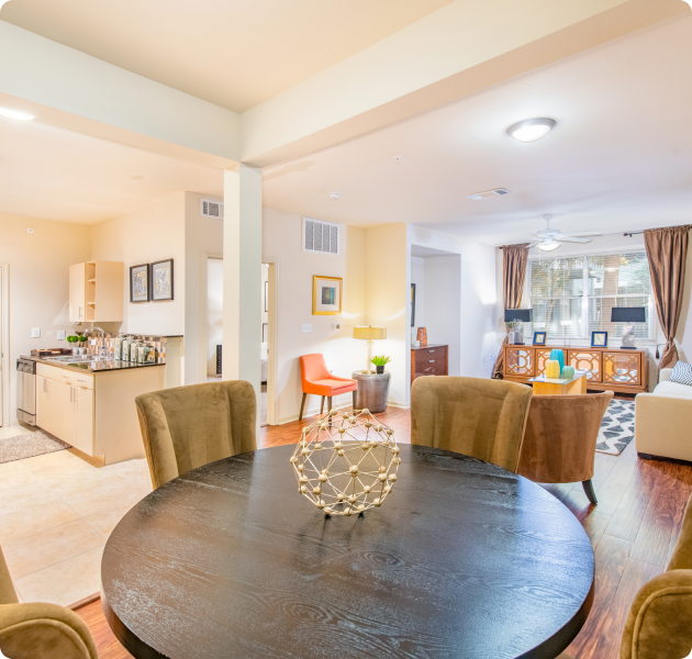 An apartment dining area with a wooden circular table and suede seats, adjacent to a living room with a couch, armchairs, and a console table near huge windows with curtains at MAA Heights luxury apartments in Dallas, TX