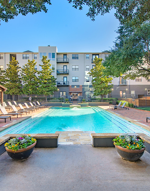A rectangular swimming pool surrounded by lounge chairs, potted plants and tall trees at MAA Heights luxury apartments in Dallas, TX