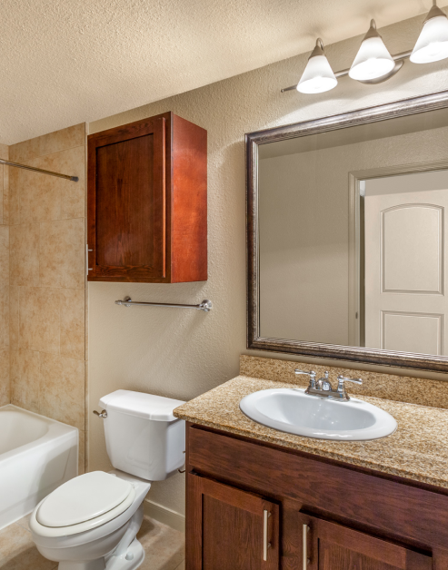 A modern bathroom featuring a bathtub, toilet, sink with a granite countertop, wooden cabinet and a large vanity mirror at MAA Heights luxury apartments in Dallas, TX