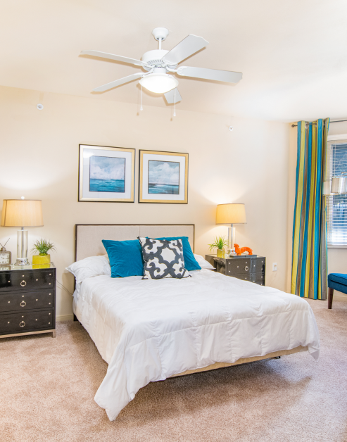 A model bedroom features a bed with white linens, blue and white decorative pillows, flanked by two dark nightstands with lamps, and a ceiling fan at MAA Heights luxury apartments in Dallas, TX