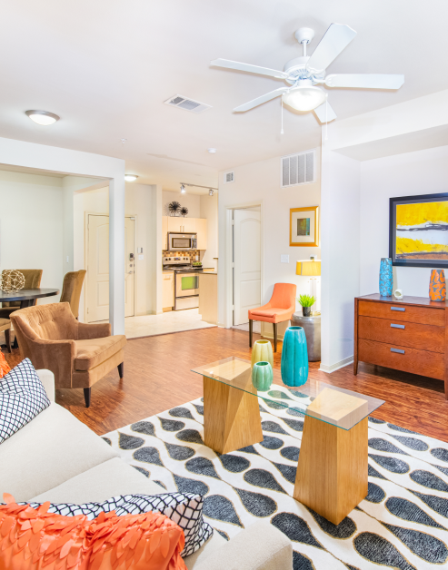 A modern living room features a white ceiling fan, a patterned rug, a light-colored sofa with orange and black cushions, and a dresser with colorful vases and wall painting, adjacent to the kitchen at MAA Heights luxury apartments in Dallas, TX