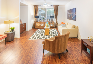 A bright living room with wood flooring, a beige sofa, an armchair, a coffee table and a patterned rug, featuring a large window with curtains at MAA Heights luxury apartments in Dallas, TX
