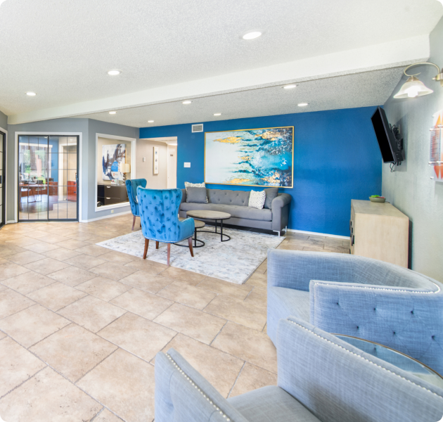 A spacious community center featuring a blue and gray couch, and a wall-mounted TV with tiled flooring at MAA Highlands North luxury apartments in Dallas, TX