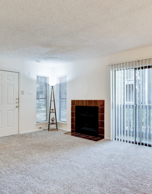 An open-space living room with a brick fireplace and side windows, carpeted floors and a stylish floor lamp at MAA Highlands North luxury apartments in Dallas, TX