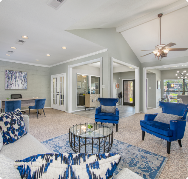 A spacious community center with a gray sofa, blue armchairs, a coffee table, and modern decor with an adjacent leasing office visible through glass doors at MAA Highwood luxury apartments in Plano, TX