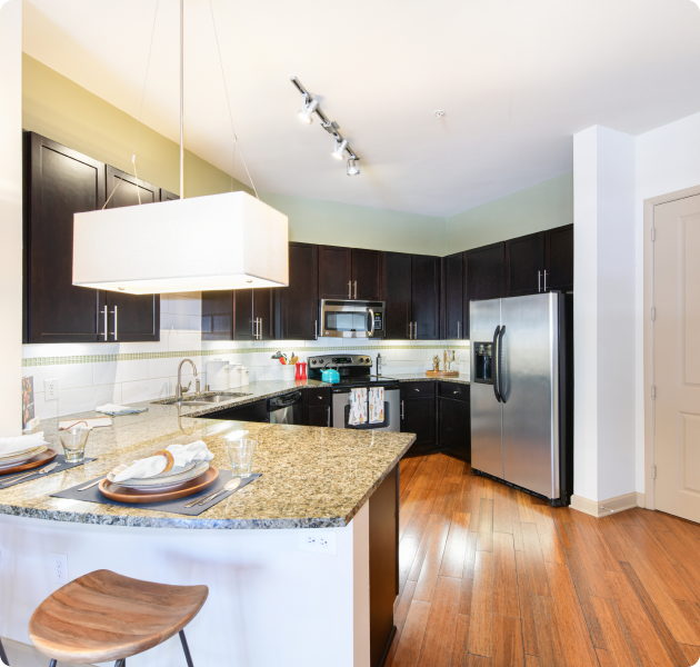 A modern apartment with a kitchen featuring stainless steel appliances including a refrigerator, granite countertop and dark wooden cabinets with hardwood flooring at MAA Katy Trail luxury apartments in Dallas, TX