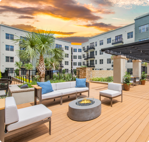 An upscale outdoor community patio area with seating around a circular fire pit on a wooden deck with palm trees, planters and a pergola at MAA Katy Trail luxury apartments in Dallas, TX