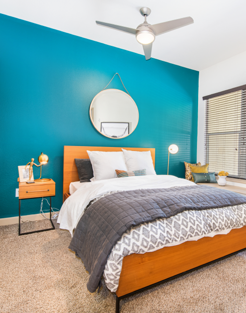 A modern bedroom with a ceiling fan and a hanging round mirror on a teal accent wall with a nightstand and a large window with blinds at MAA Katy Trail luxury apartments in Dallas, TX