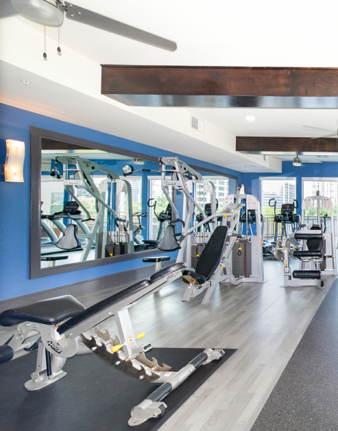 A luxurious fitness center featuring an exercise bench next to a large wall mirror, ceiling fans and various workout equipment at MAA Katy Trail luxury apartments in Dallas, TX
