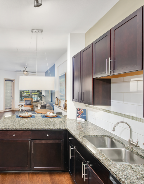 A luxurious kitchen featuring a granite countertop with dark wooden cabinets, stylish wall-mounted lights and wood floors at MAA Katy Trail luxury apartments in Dallas, TX