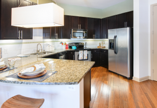 A modern kitchen with stainless steel appliances including a refrigerator, dark wood cabinets with granite countertops and hardwood flooring at MAA Katy Trail luxury apartments in Dallas, TX
