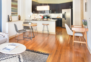 A modern living room with a white coffee table and wooden writing desk next to an open kitchen with a small dining area and wood floors at MAA Katy Trail luxury apartments in Dallas, TX