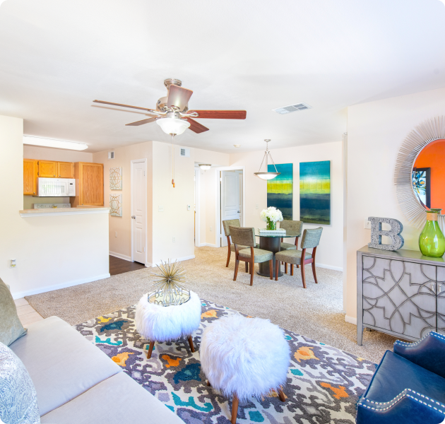 An apartment living area with a ceiling fan and couch sitting area with a round mirror above an accent cabinet next to a dining area with hanging light fixture and wall artwork next to the kitchen at MAA Los Rios luxury apartments in Plano, TX