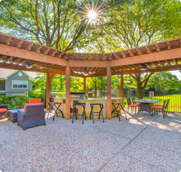 A large gated outdoor patio area covered with a wooden pergola and ceiling fans, featuring a grill station with barstools, tables with chairs and lounge seating area surrounded by trees and grass at MAA Los Rios luxury apartments in Plano, TX