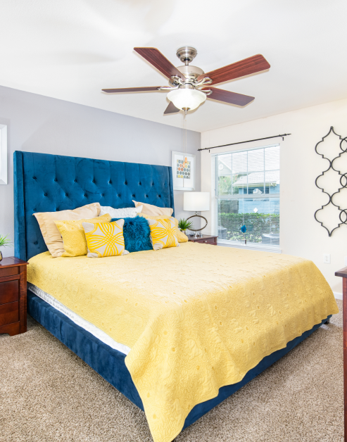 A modern bedroom with a ceiling fan and a nightstand with a lamp next to bed with blue tufted headboard at MAA Los Rios luxury apartments in Plano, TX