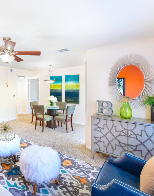 A vibrant living room with stylish round mirror above an accent cabinet, a blue armchair next to dining area with ceiling fan over a dining table with wall paintings at MAA Los Rios luxury apartments in Plano, TX