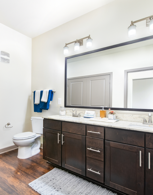 Modern bathroom with a large vanity mirror, white granite countertop with dark brown cabinets and wood floors at MAA Market Center luxury apartments in Plano, TX