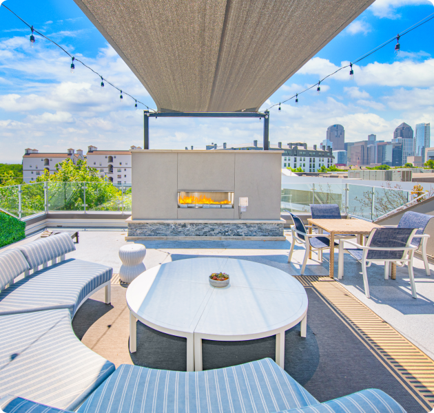 A luxurious rooftop covered community lounge featuring a fireplace next to patio table and chairs, and a large round sitting area with a round white center table at MAA McKinney Ave luxury apartments in Dallas, TX