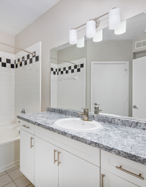 Spacious upscale bathroom with light-colored cabinets, stylish granite countertop and large vanity mirror next to bathtub and shower at MAA McKinney Ave luxury apartments in Dallas, TX