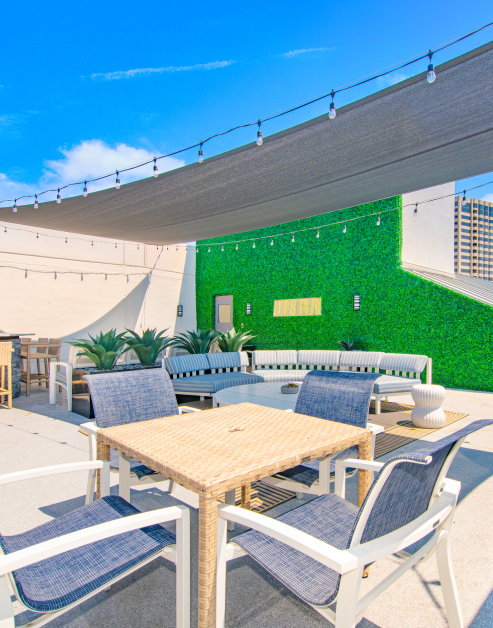 An open rooftop patio featuring a table and chairs with couch seating enhanced by an artificial grass wall backdrop with a canopy decorated with market lights at MAA McKinney Ave luxury apartments in Dallas, TX