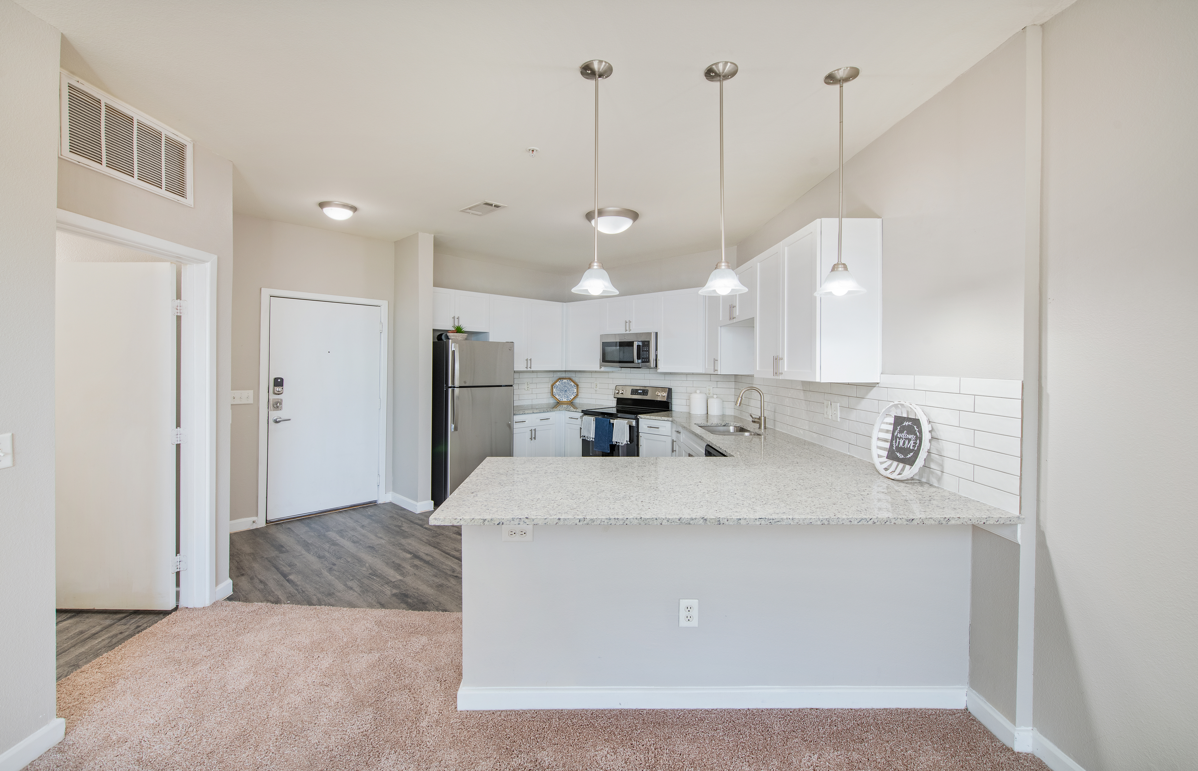 A side view of an upscale kitchen with hanging light fixtures, a granite countertop with white cabinets and hardwood floors connected to living room at MAA Medical District luxury apartments in Dallas, TX