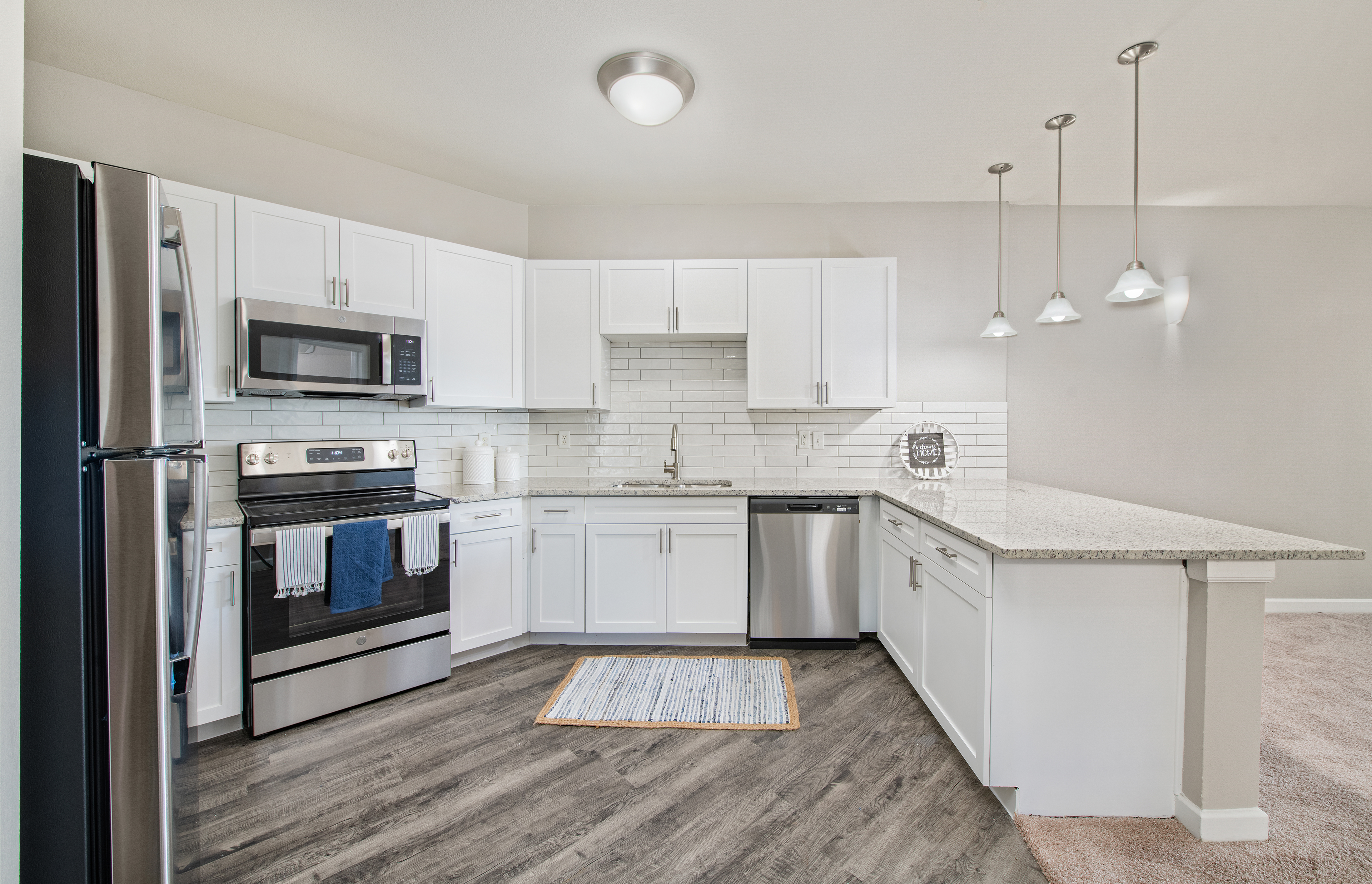 A side view of an open-space kitchen with hanging light fixtures and white cabinets with tiled backsplash, granite countertop, and hardwood flooring connected to a living room at MAA Medical District luxury apartments in Dallas, TX