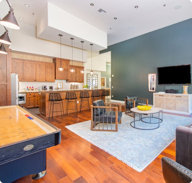 A spacious community lounge area with a wall-mounted TV next to seating area with armchairs, an open kitchen with stylish lights and barstools and air hockey table at MAA Medical District luxury apartments in Dallas, TX