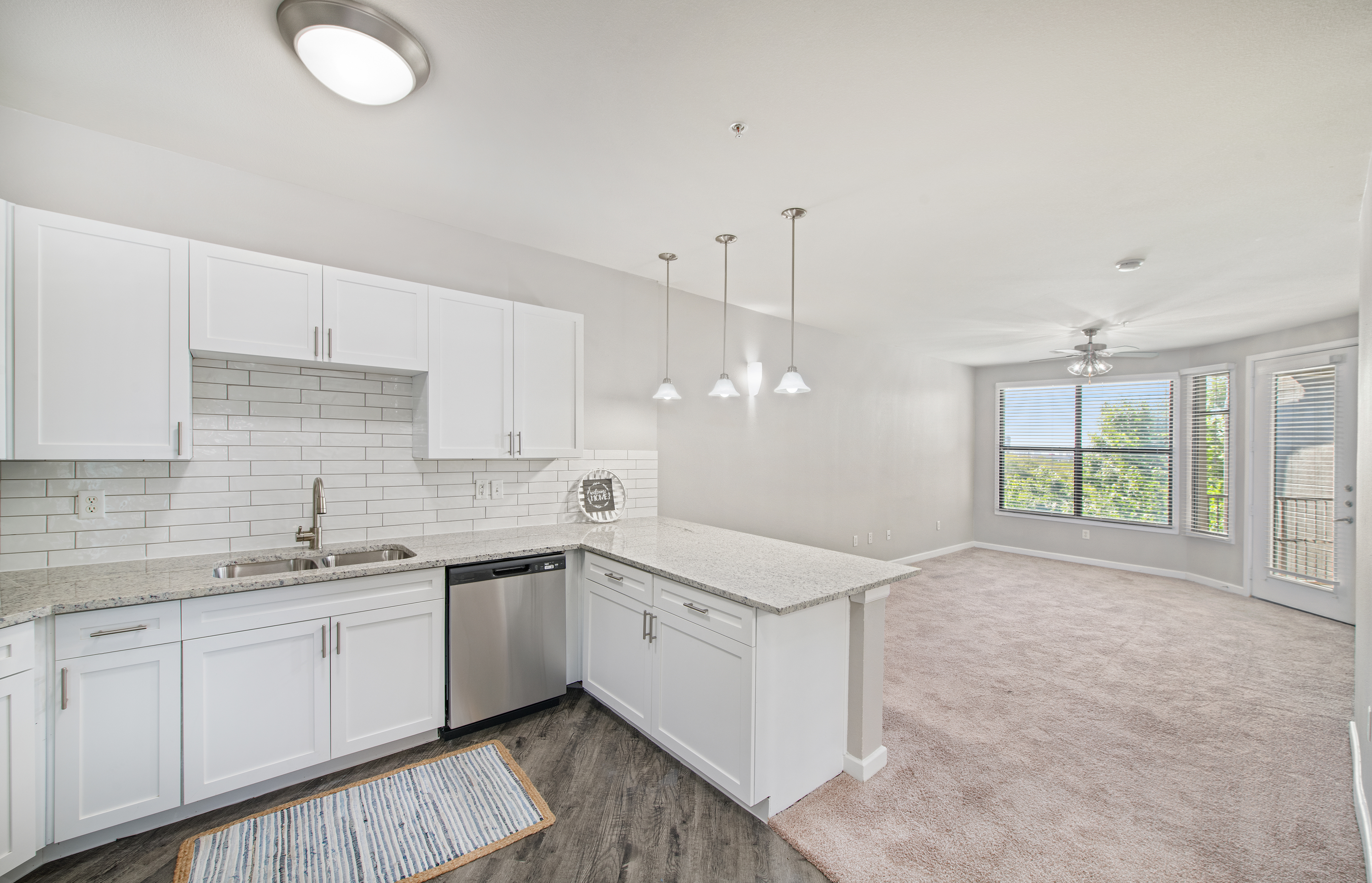 A side view of an open-space kitchen with hanging light fixtures and white cabinets with tiled backsplash, granite countertop, and hardwood flooring connected to a living room at MAA Medical District luxury apartments in Dallas, TX