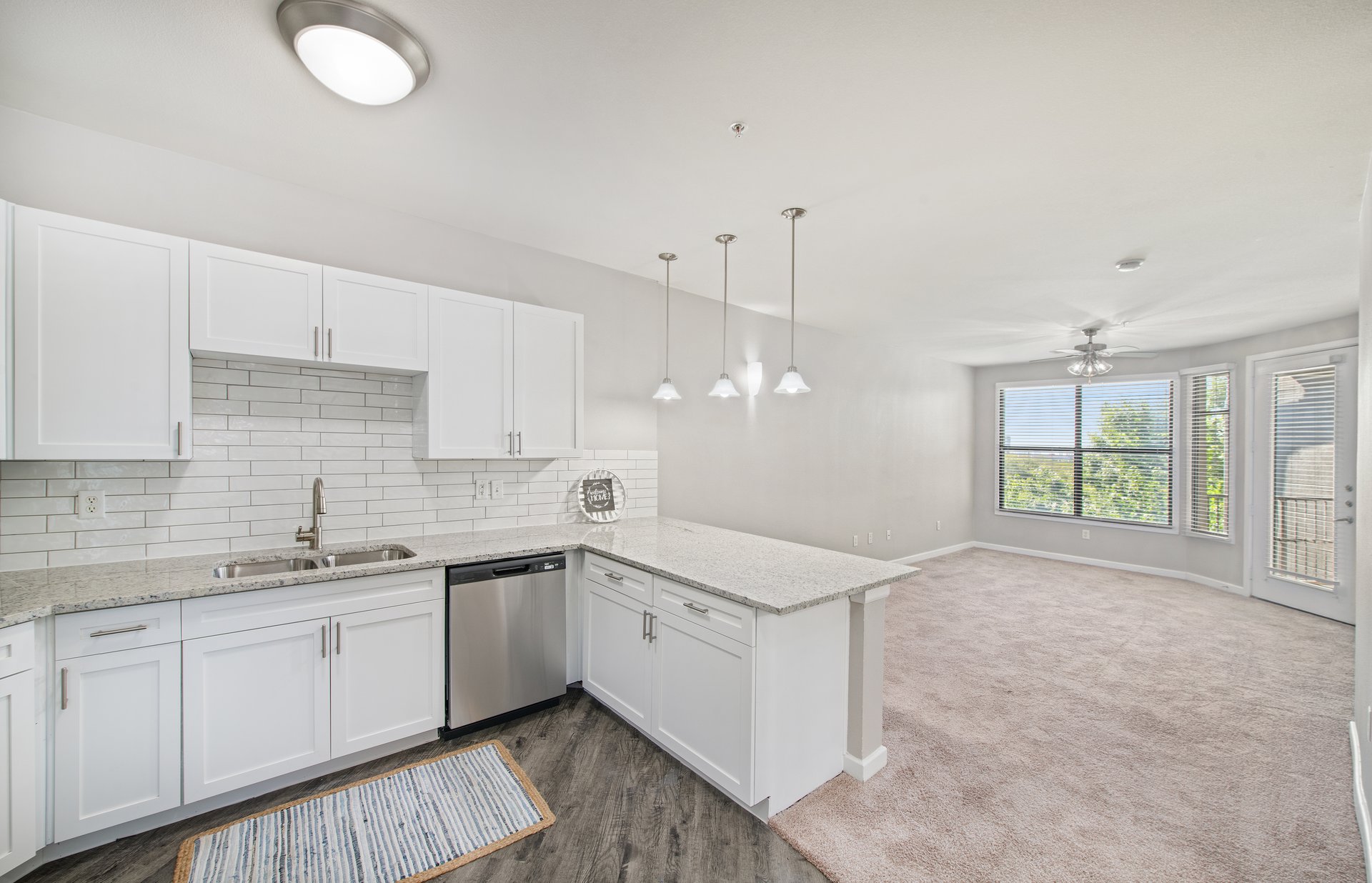 A side view of an open-space kitchen with hanging light fixtures and white cabinets with tiled backsplash, granite countertop, and hardwood flooring connected to a living room at MAA Medical District luxury apartments in Dallas, TX