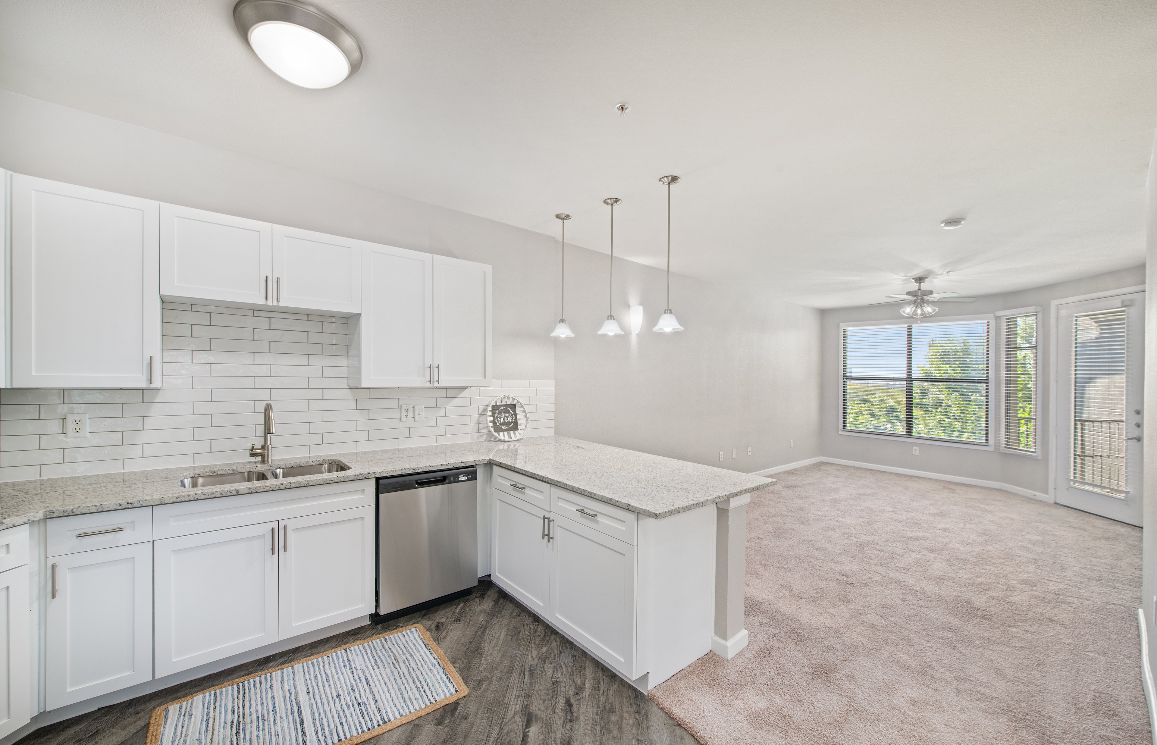 A side view of an open-space kitchen with hanging light fixtures and white cabinets with tiled backsplash, granite countertop, and hardwood flooring connected to a living room at MAA Medical District luxury apartments in Dallas, TX