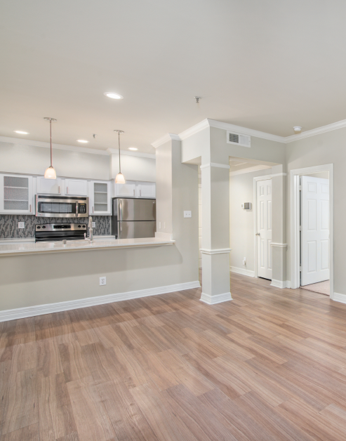 A large, vacant living room with rich hardwood flooring, adjacent to the kitchen featuring ceiling light fixtures above the bar top counter at MAA Meridian luxury apartments in Dallas, TX