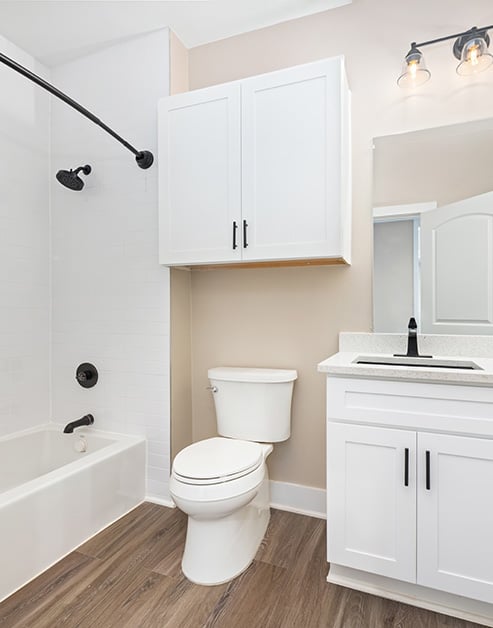 Bathroom with single sink vanity with white cabinets, a wall mounted white cabinet above toilet next to a tiled bathtub shower with wood floors at MAA North Hall luxury apartments in Dallas, TX