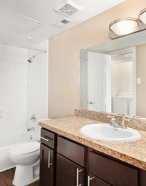 A modern bathroom featuring a granite countertop with dark wood cabinets next to a doorway to bathtub and wood floors at MAA Remington Hills luxury apartments in Irving, TX